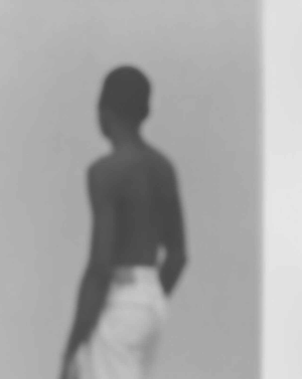 Blurred silhouette of a shirtless figure standing against a light gray wall, rendered in soft black and white tones. The abstracted form and ethereal quality suggest movement or a fleeting moment captured through deliberate focus techniques.