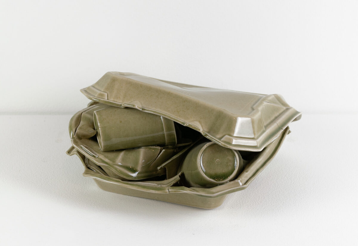 A pale celadon-glazed porcelain sculpture depicting an open takeout container with crumpled paper napkins and two cups nestled inside, rendered in refined ceramic material that mimics the appearance of disposable styrofoam packaging.
