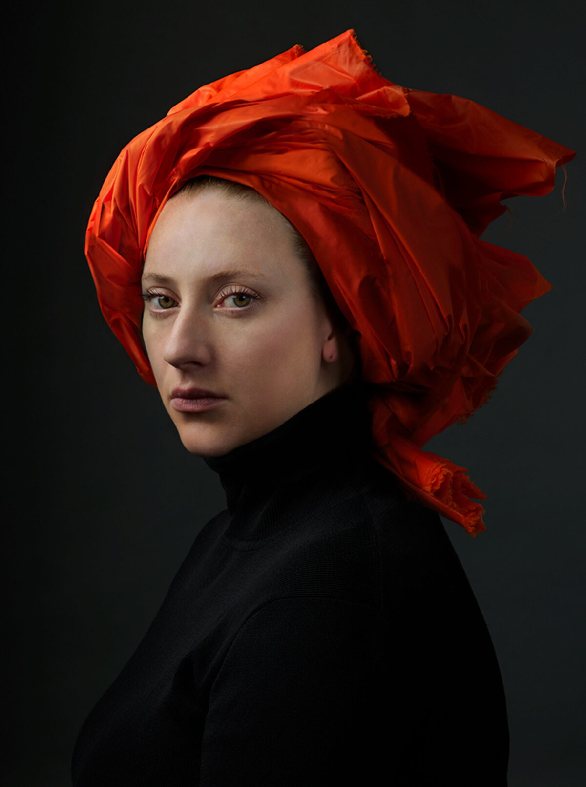 Portrait of a person with a pale complexion wearing a voluminous bright red-orange fabric wrapped around their head, set against a dark gray background. The figure is dressed in black and gazes toward the viewer in three-quarter profile, creating a striking contrast between the vivid headwrap and subdued tones of the clothing and background.