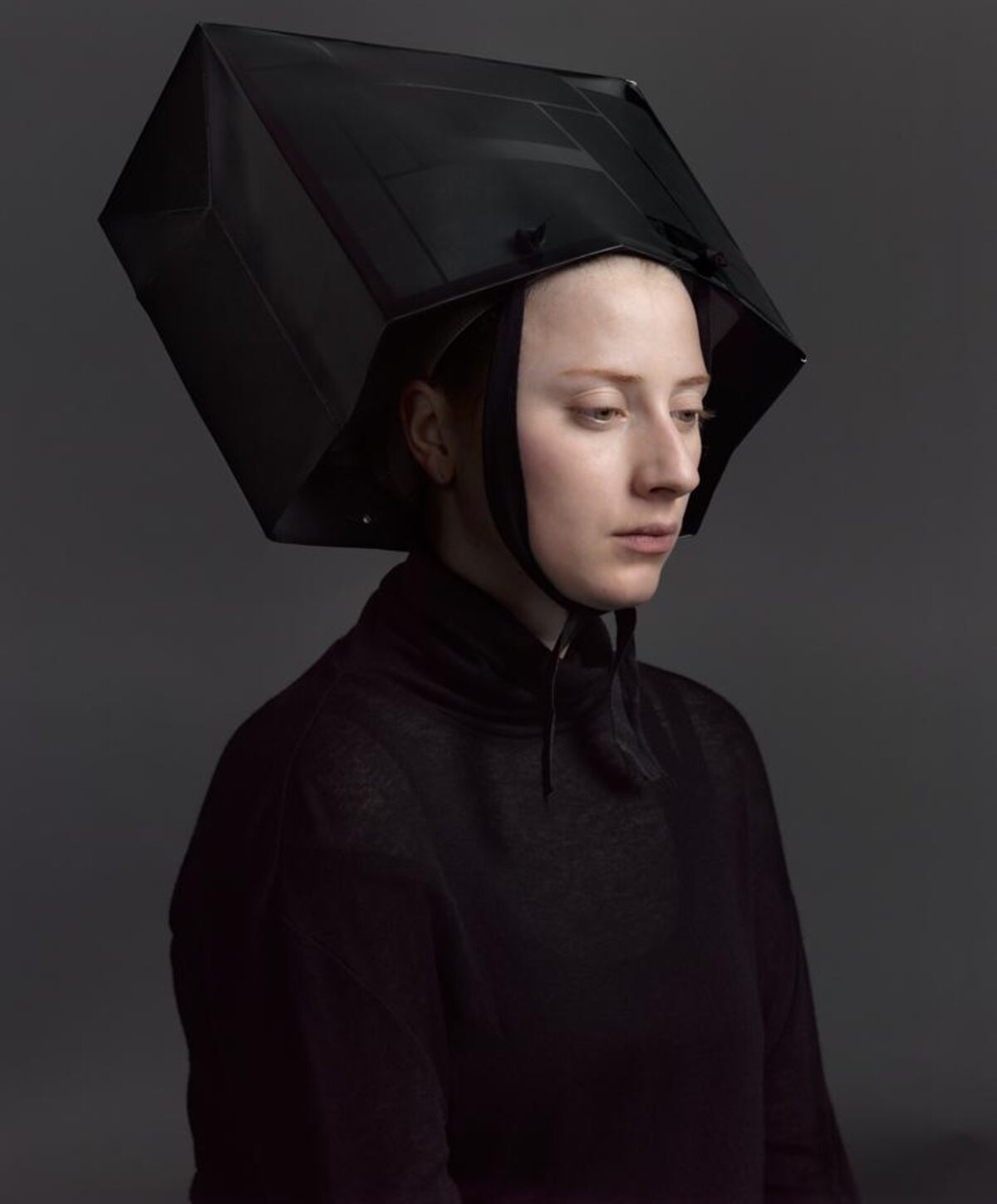 A figure in profile wearing a dark, high-necked garment with a striking geometric black hood that extends outward in angular, box-like planes. The minimalist composition is set against a neutral gray background, emphasizing the sculptural quality of the headpiece and the contemplative downward gaze of the pale-faced subject.