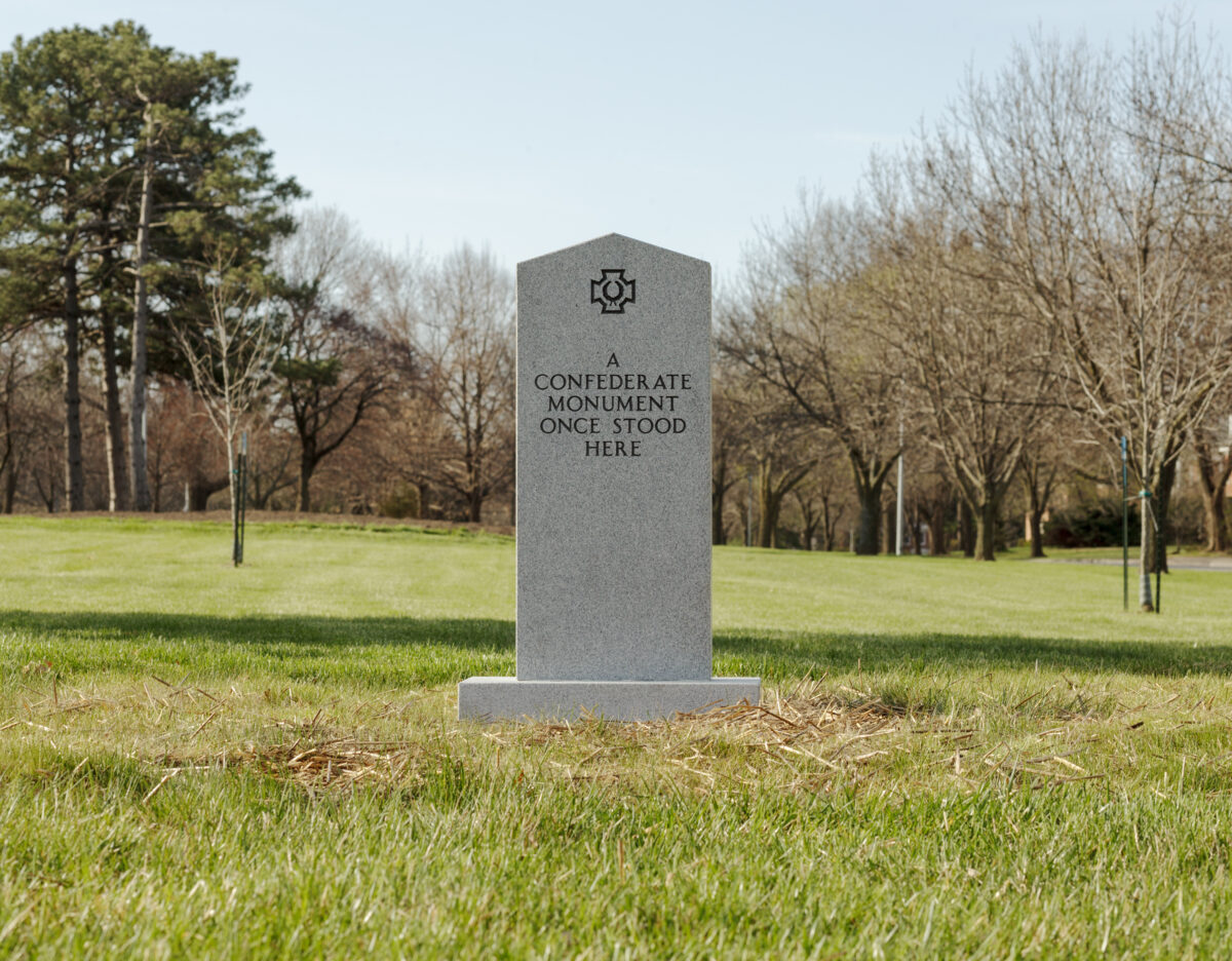 A gray stone monument stands alone in a grassy park surrounded by bare and evergreen trees. The obelisk-shaped marker bears an engraved symbol at its top and text reading "A CONFEDERATE MONUMENT ONCE STOOD HERE."
