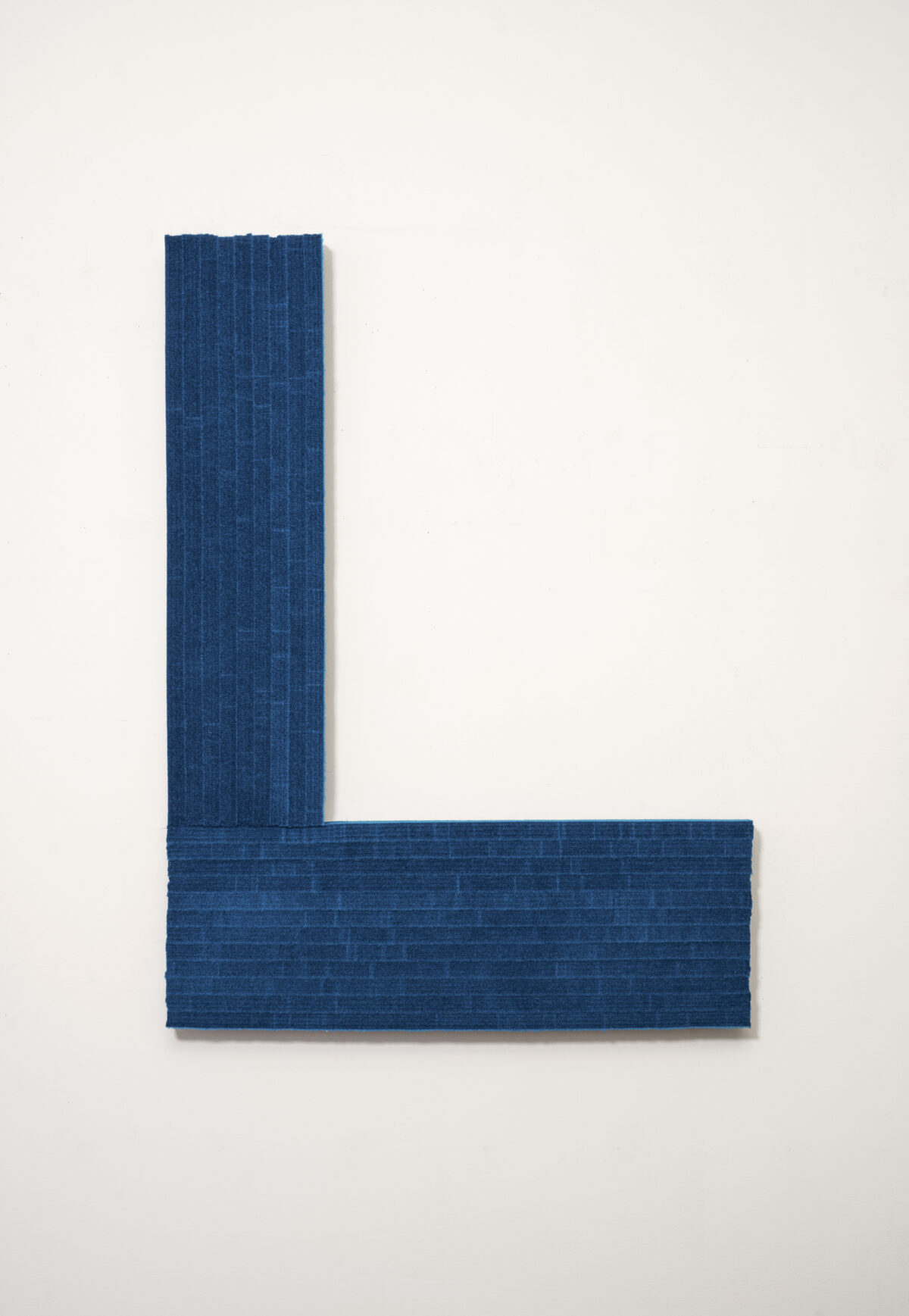 A deep blue textile panel arranged in an L-shaped composition against a white wall. The material has a subtle woven texture with visible vertical striations, creating a minimalist geometric form.