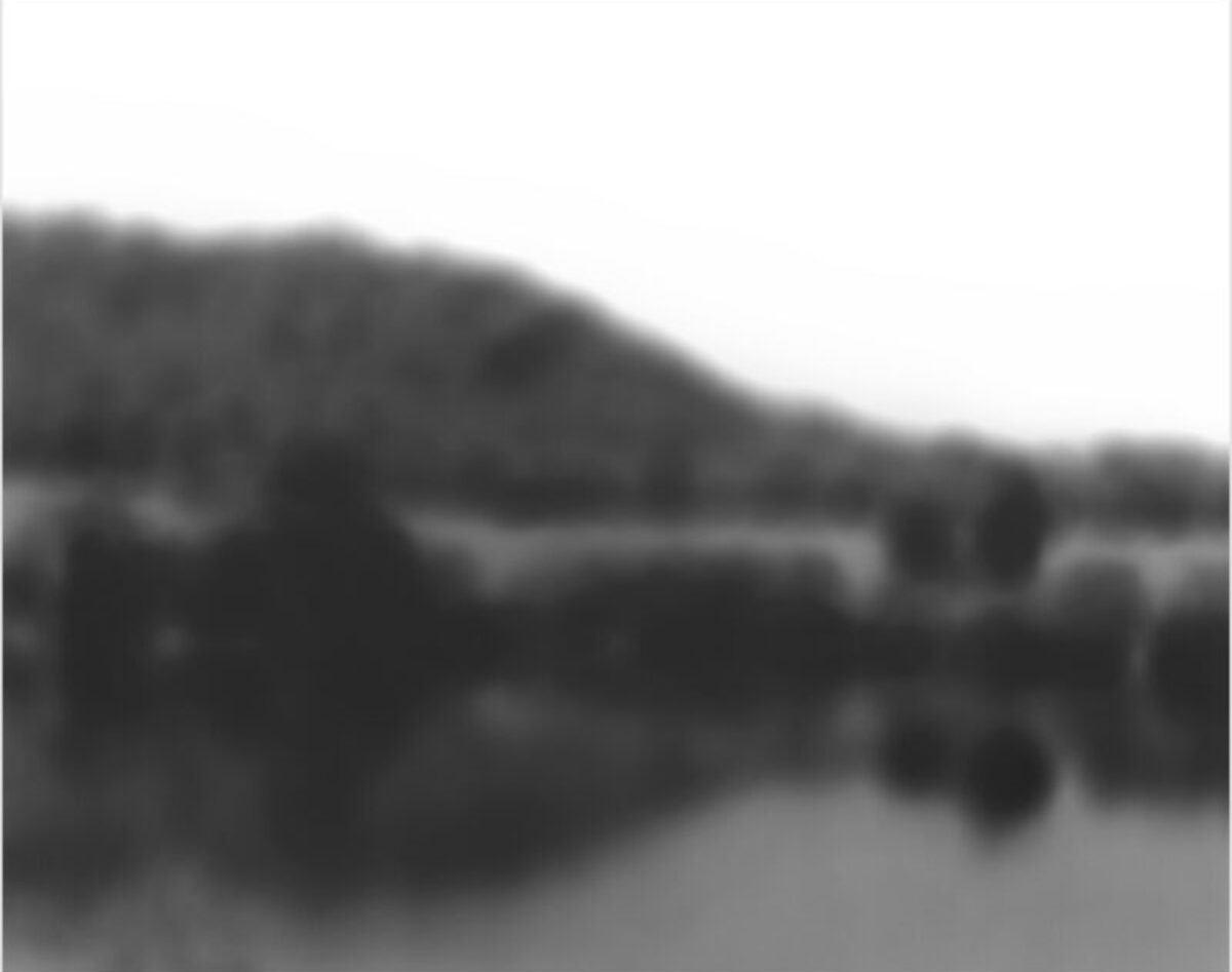 Softly focused black and white landscape depicting a body of water with forested hills or mountains rising beyond it, rendered in subtle tonal gradations that emphasize atmospheric depth and the ephemeral quality of the scene.