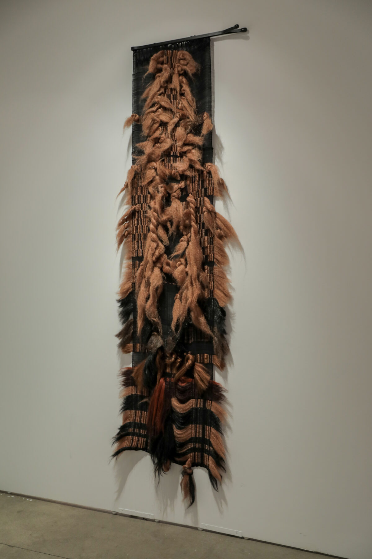 A vertical tapestry featuring expressive carved or sculpted faces in warm brown tones mounted on dark fabric, with synthetic hair and natural fibers extending outward along the sides, creating a textured, three-dimensional effect against a white gallery wall.