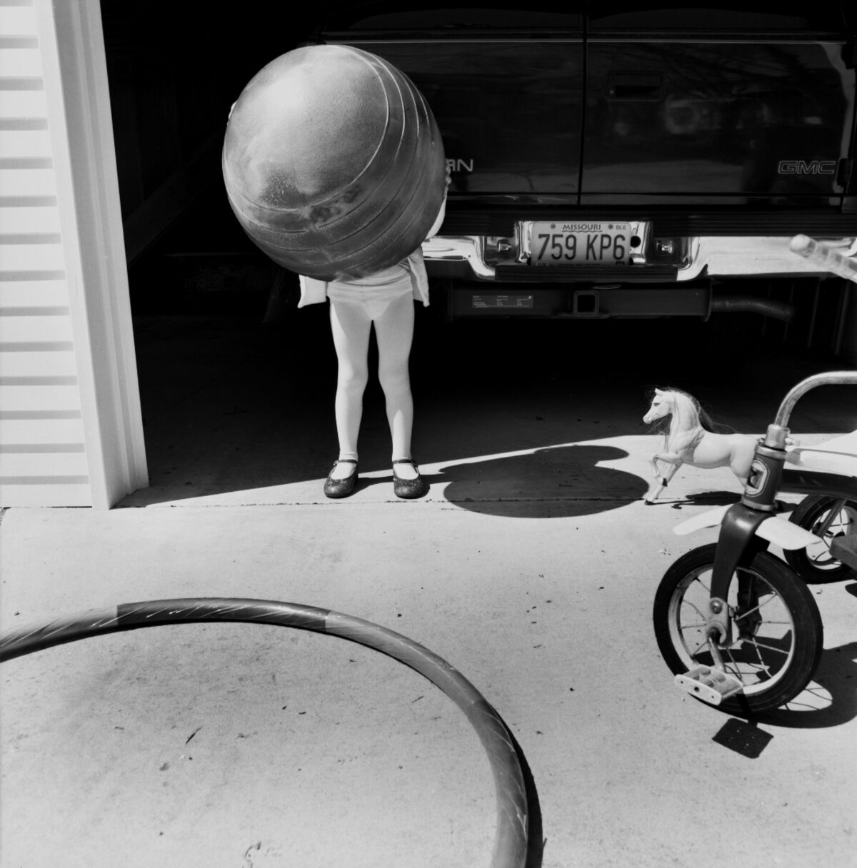 Black and white photograph of a person holding an enormous striped ball that obscures their head and upper body, standing in a driveway beside a GMC truck with visible license plate and a child's tricycle. A small toy horse figure sits nearby on the concrete pavement.