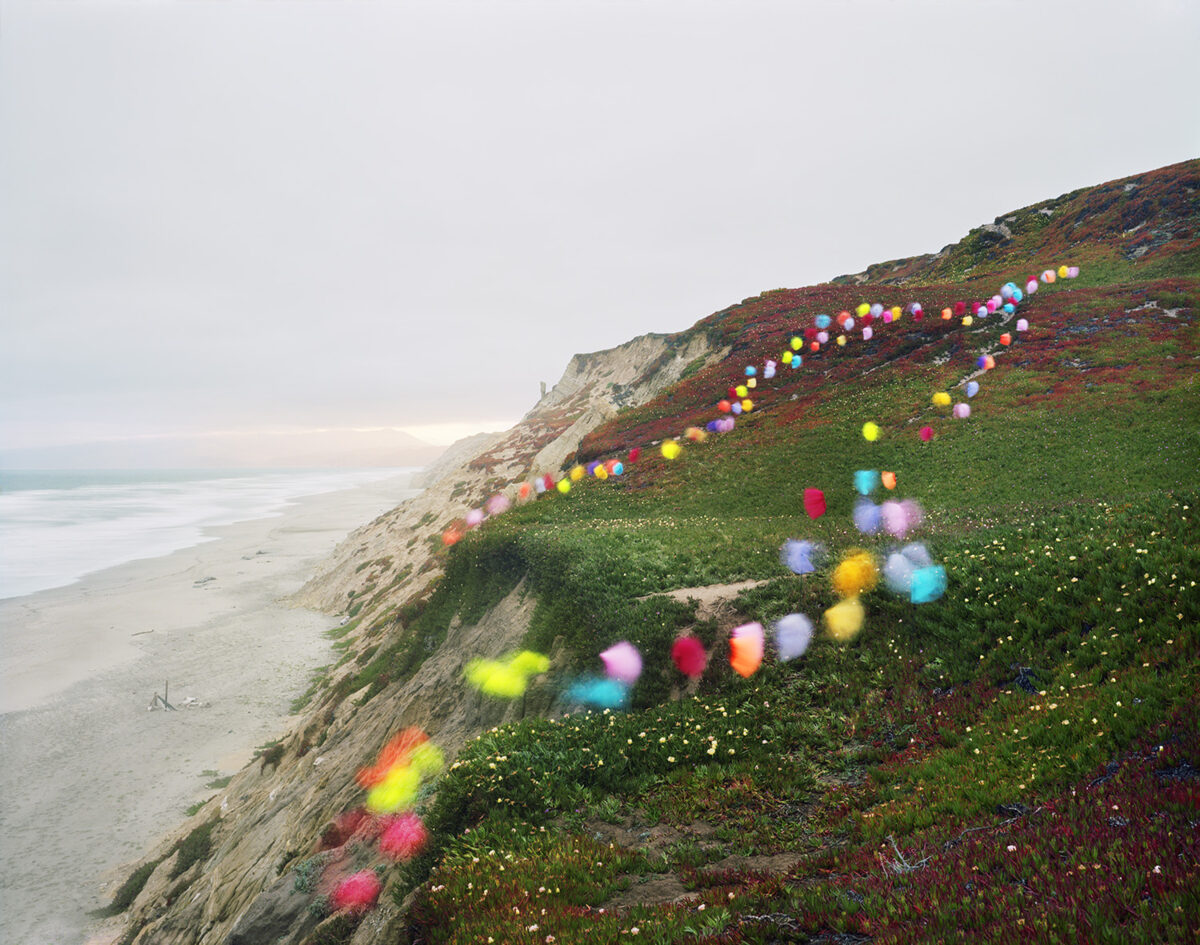 Colorful inflated spheres in shades of pink, yellow, blue, cyan, and red are arranged in curved lines across a coastal hillside covered in green vegetation and rust-colored plants. The spheres create a playful path descending toward a dramatic eroded cliff face and sandy beach below, with calm ocean visible in the distance.