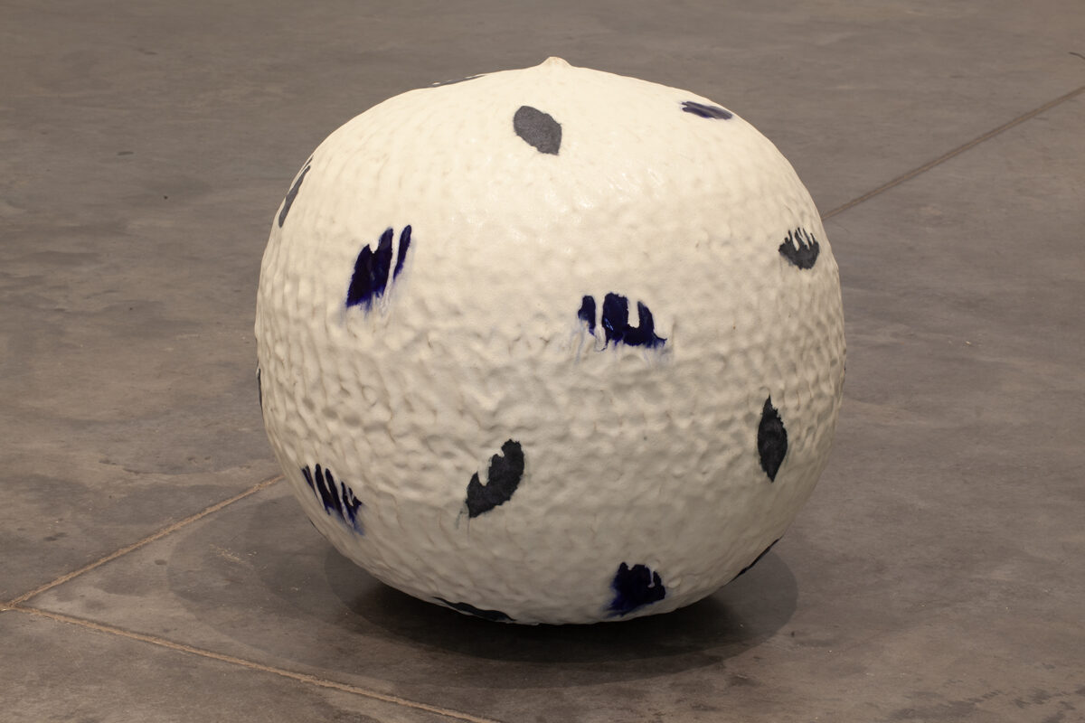 A large cream-colored spherical ceramic form sits on a concrete floor, its surface marked with gestural brushstrokes in deep blue and charcoal gray that resemble organic marks or leaf impressions distributed across the rounded form.