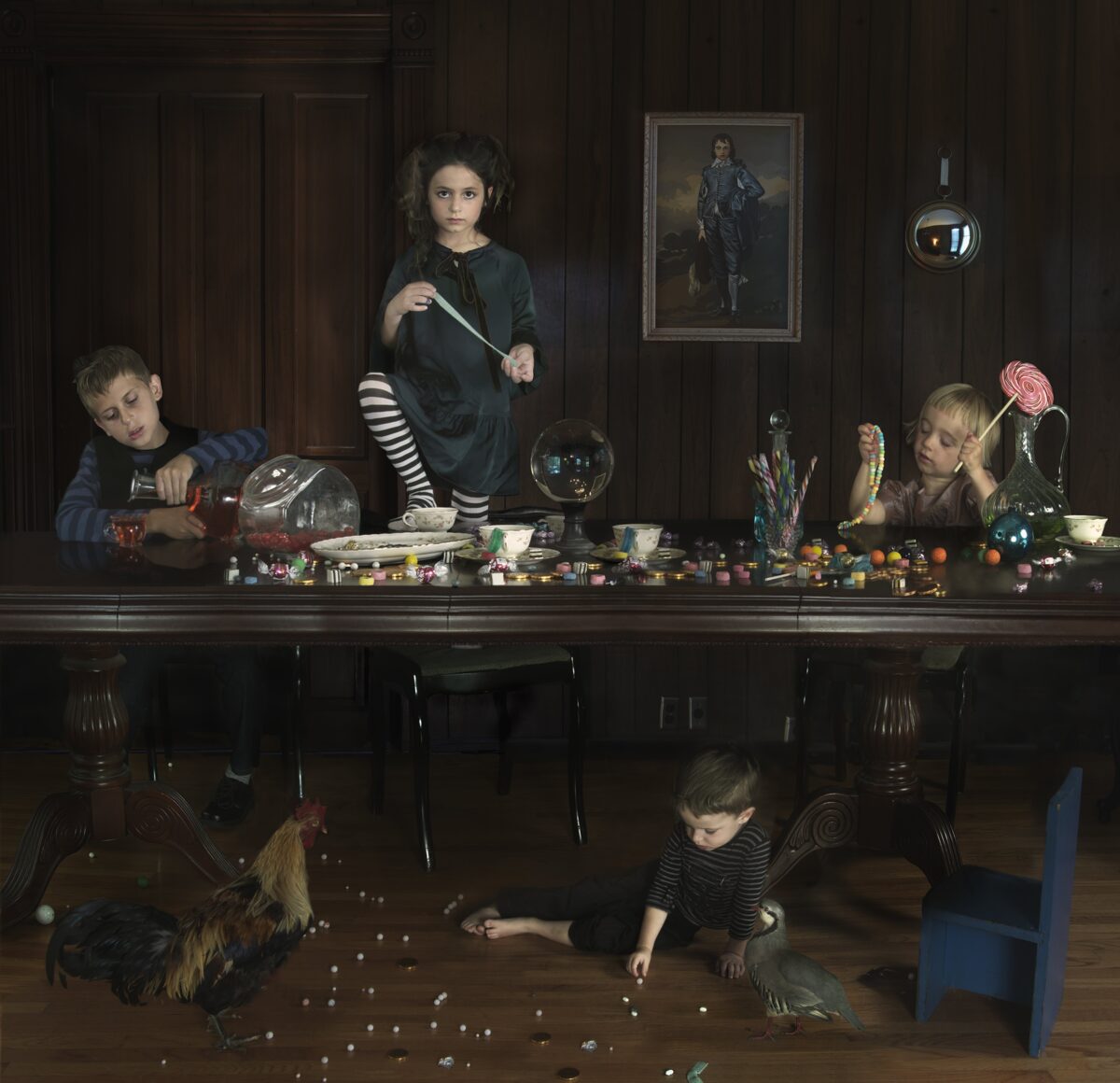 Children of varying ages are arranged at a long dark table laden with candies and toys, with two younger children and a rooster on the floor below scattering small round objects. The scene is set in a wood-paneled interior with framed portraits on the walls, rendered in rich, dark tones with carefully controlled lighting that emphasizes the children's faces and the colorful items scattered across the surfaces.