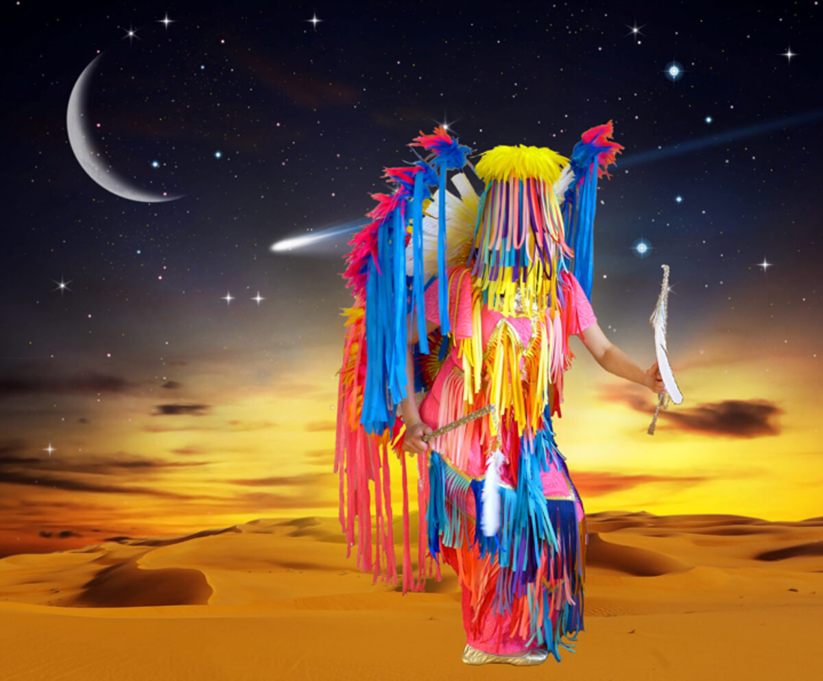 A vibrant, colorful figure stands in a desert landscape beneath a starlit night sky with a crescent moon and comet. The figure is draped in flowing ribbons and fringe in shades of hot pink, bright yellow, electric blue, and red, creating an explosive, dynamic silhouette against the golden sand and warm sunset horizon.