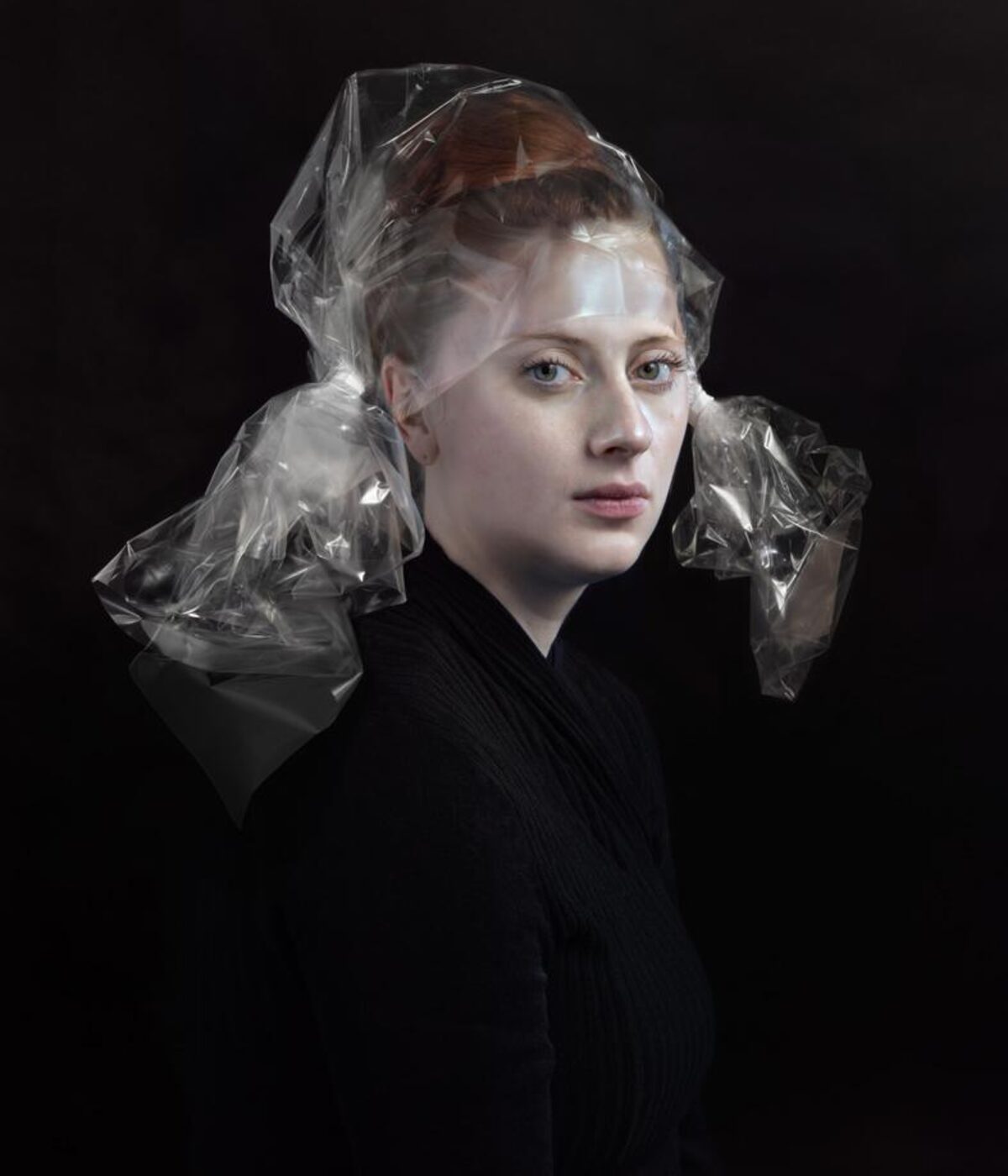 Portrait of a fair-skinned woman with reddish-brown hair styled close to her head, wearing a black ribbed garment against a black background. Transparent cellophane material is draped over her head and shoulders, creating translucent folds and reflective surfaces that frame her face and extend down her back.