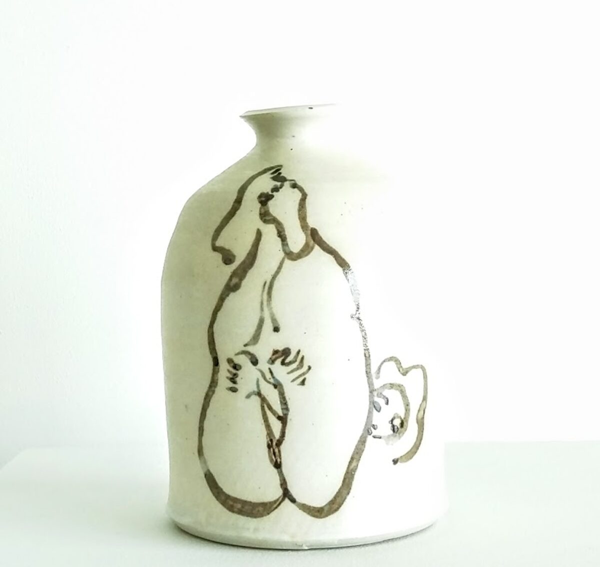 A cream-colored ceramic vessel with a narrow neck and rounded body features a minimalist ink drawing of a reclining nude figure rendered in loose, gestural lines. A small handle extends from the lower right side of the form.
