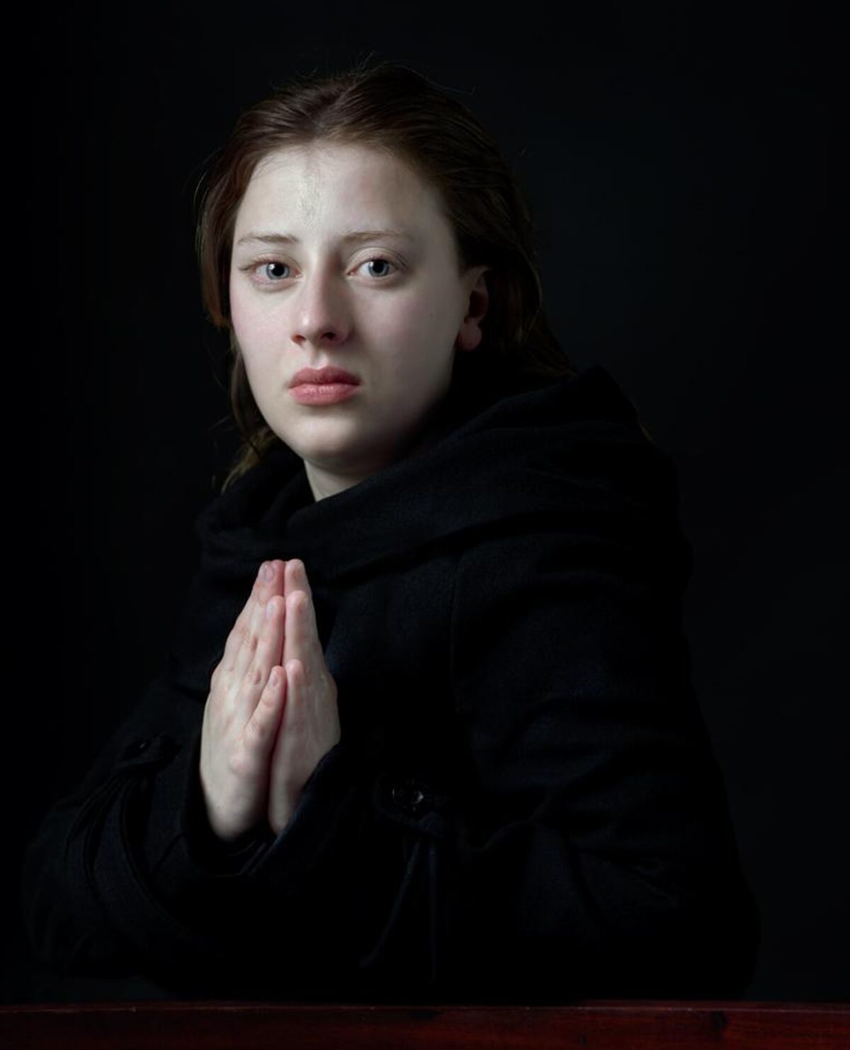 Portrait of a young woman with dark hair against a black background, wearing a black garment with her hand raised to her chest in a contemplative gesture. The stark lighting emphasizes her pale complexion and direct gaze toward the viewer, creating an intimate, introspective mood.