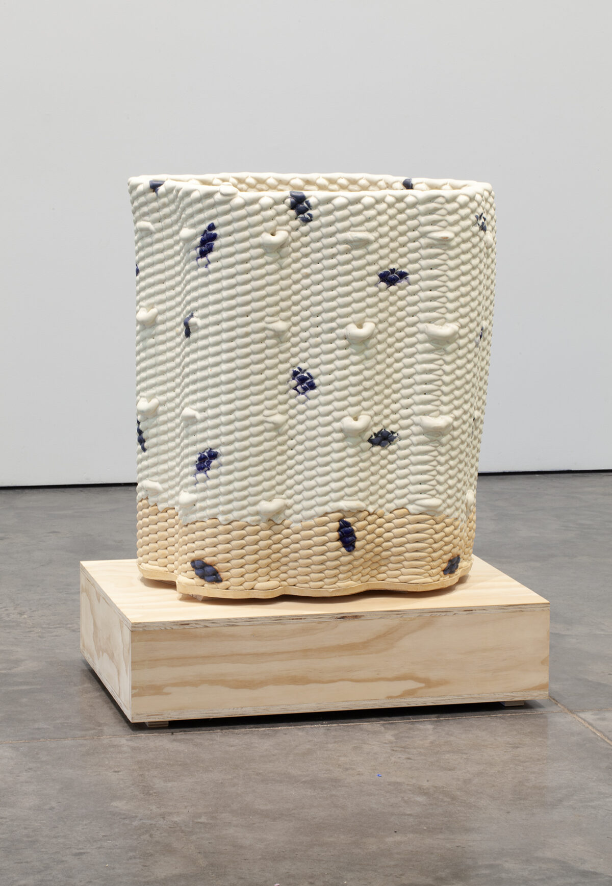 A cylindrical ceramic vessel with a textured, bumped surface in cream and pale yellow tones, scattered with dark blue glazed marks. The piece sits on a light wooden base against a gallery floor and white wall.