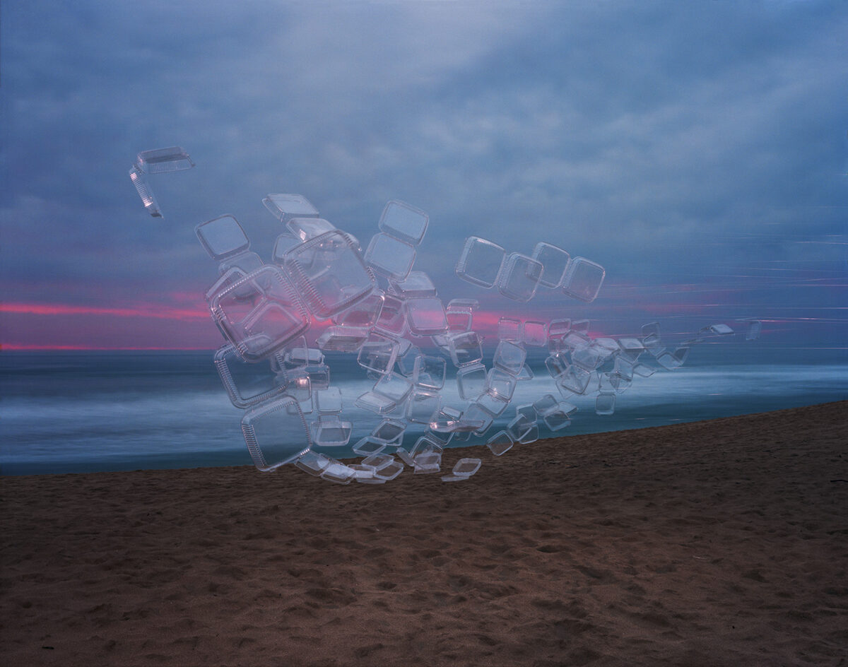 Translucent plastic takeout containers arranged in a sculptural pile on a beach at dusk, glowing with pink and blue light against a twilight sky and calm ocean horizon.