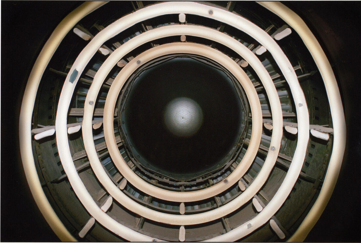 Concentric circles of cream and white rings with metal fixtures frame a dark central aperture, creating a geometric spiral effect. The composition suggests looking down into a cylindrical industrial or architectural structure, with alternating bands of light and shadow receding toward a barely visible illuminated center.