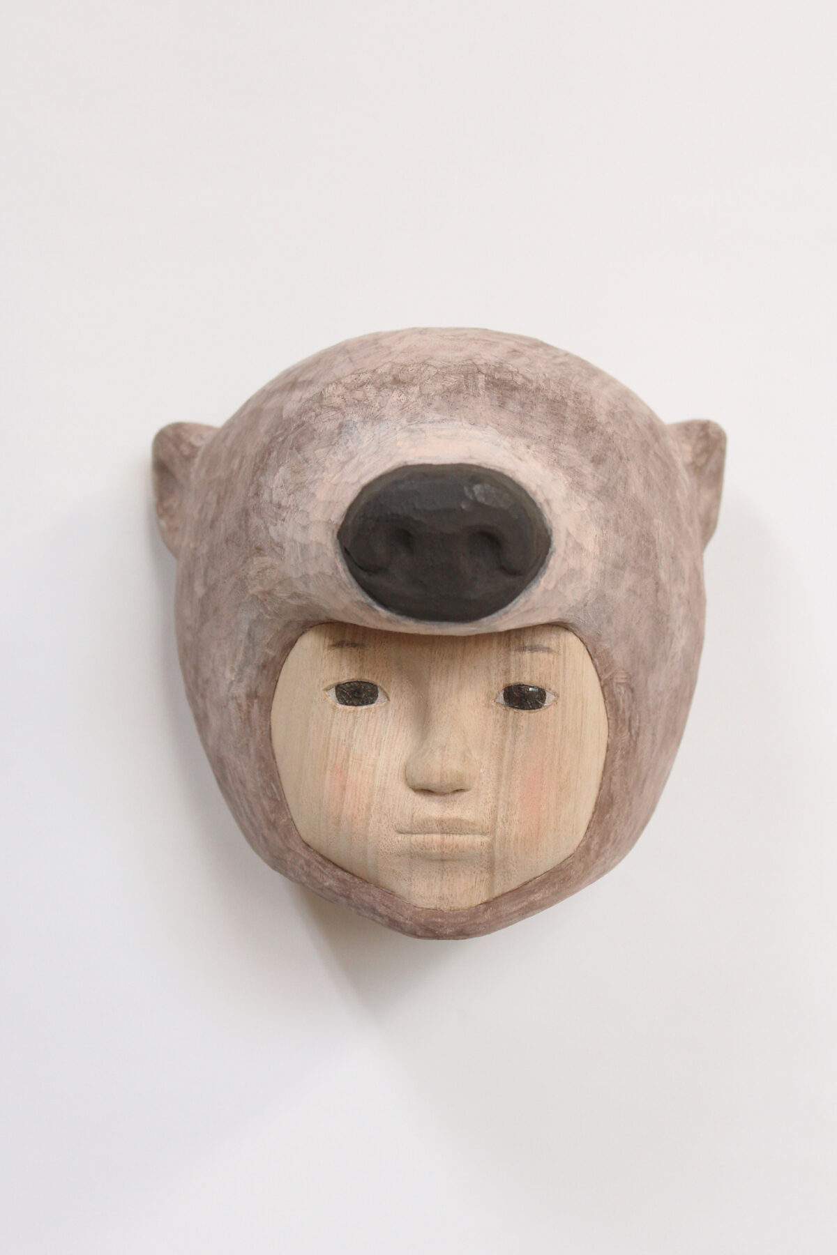 Carved wooden mask depicting a human face with closed eyes and neutral expression, framed by a rounded animal head with pointed ears and a large dark oval opening where the eyes would be. The lighter wood face contrasts with the darker weathered tones of the surrounding animal form.