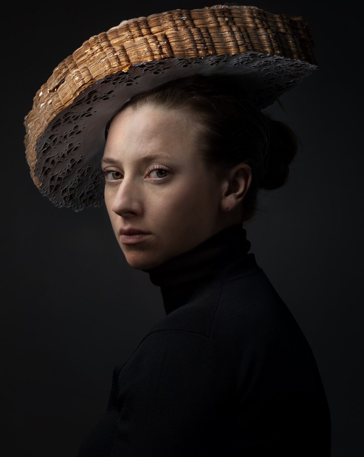 Portrait of a woman in profile against a dark background, wearing a black turtleneck and an elaborate woven hat with a straw-colored crown and dark perforated brim that extends dramatically to the side.