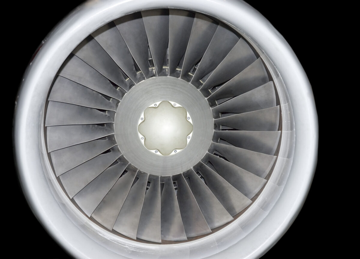 Front-facing view of a jet engine turbine with concentric metal blades radiating from a central hub, rendered in grayscale tones against a black background. The precise, symmetrical composition emphasizes the industrial geometry and reflective surfaces of the machinery.