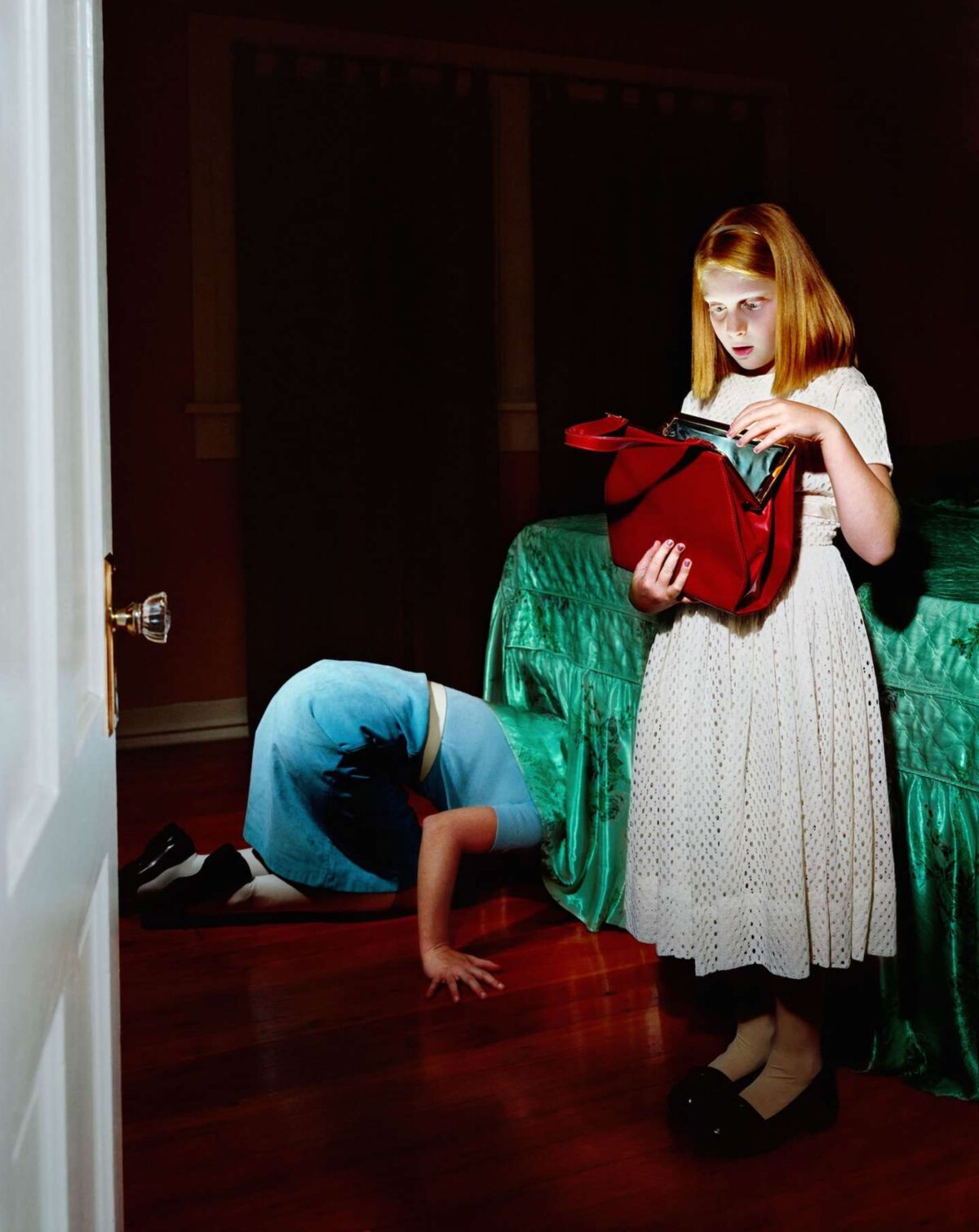 A young girl with reddish-blonde hair stands in a darkened interior space wearing a white eyelet dress, holding an open red purse. Behind her, a person in blue jeans bends forward on a green quilted fabric, their head bowed out of frame. The composition uses dramatic lighting and rich jewel tones against deep shadows.