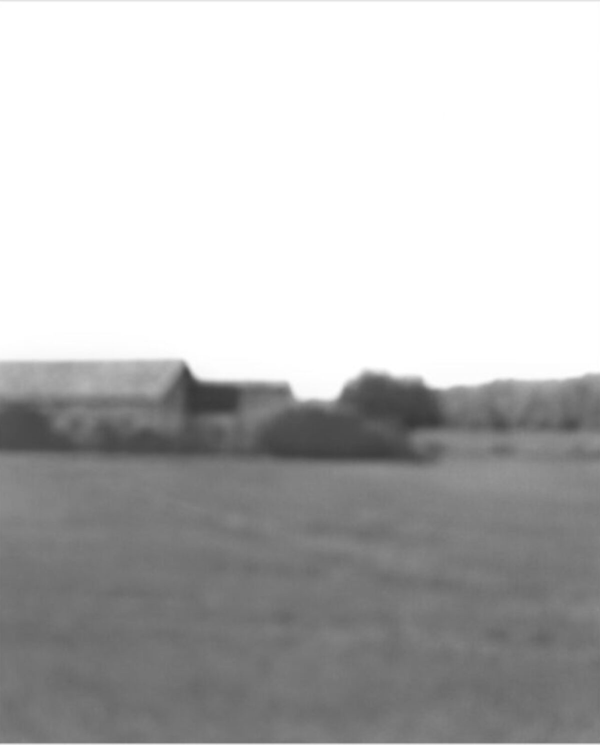 Monochromatic landscape photograph featuring a distant rural scene with farm buildings and trees along the horizon, rendered in soft focus with a hazy, atmospheric quality. The composition is dominated by an empty foreground of flat terrain beneath a pale sky.
