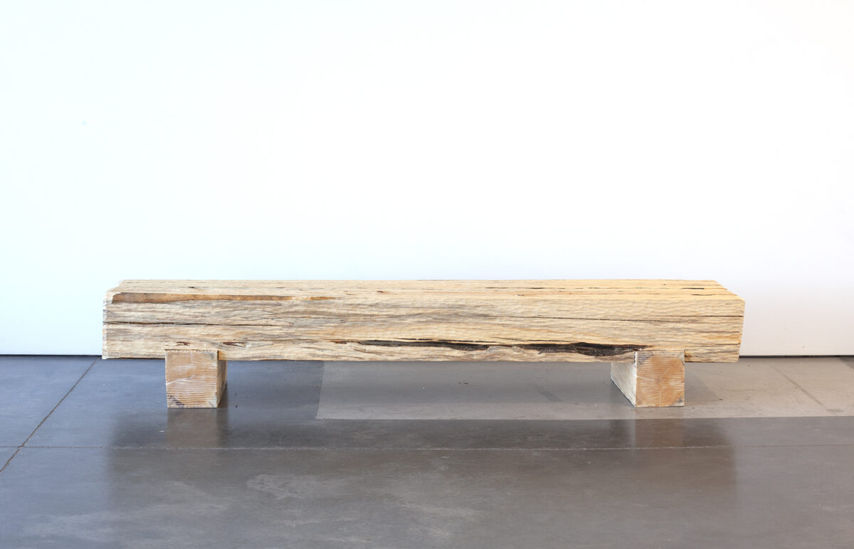 A long wooden bench made from a single weathered log with a dark burn scar running across its top surface, supported by two rectangular wooden blocks, sits on a polished concrete floor against a white wall.
