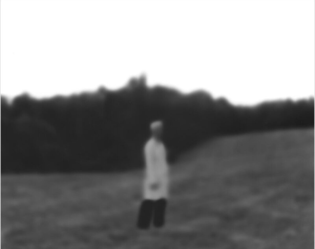 A solitary figure in a white shirt and dark pants stands in a vast, empty field, facing away from the camera toward a dark treeline on the horizon. The black and white photograph has a soft, slightly out-of-focus quality that emphasizes the isolation of the lone person in the expansive landscape.