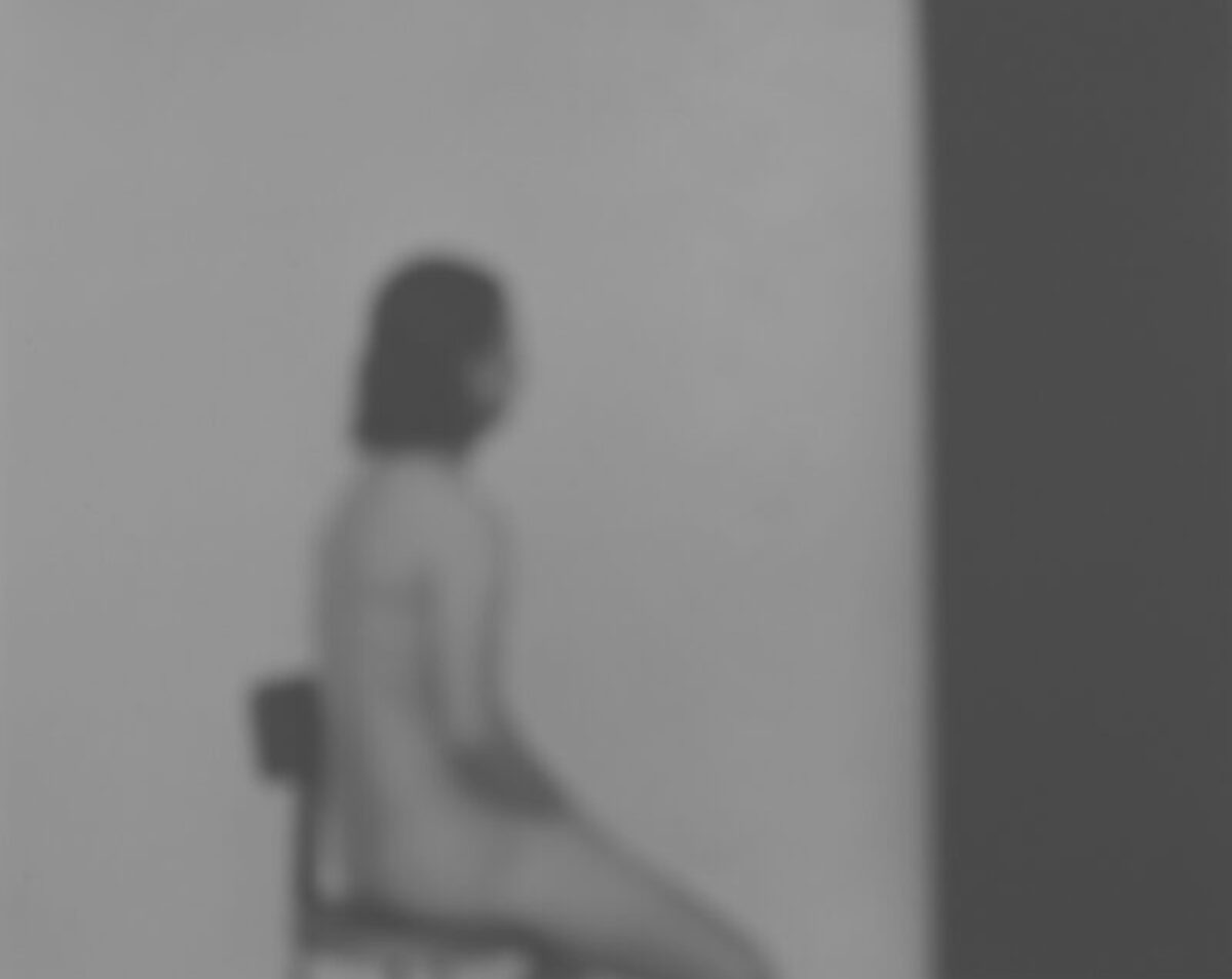 A blurred figure with dark hair sits in profile against a light wall, rendered in soft grayscale tones. The photograph's subtle focus and muted palette create an introspective, contemplative mood.