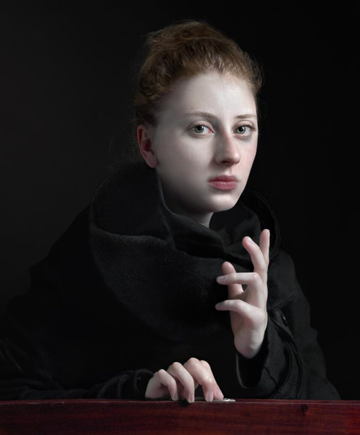A young woman with reddish-brown hair styled upward gazes toward the camera against a dark background. She wears a black garment and rests her pale hands on a dark wooden surface, her expression contemplative and direct.