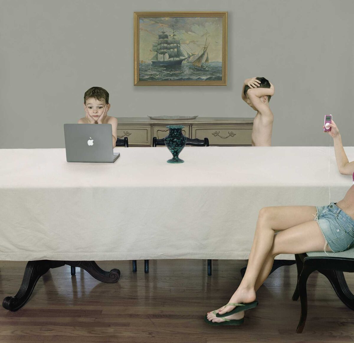 A surreal domestic scene depicts three disembodied figures positioned at a white table against a gray wall. On the left, a young boy's head rests on his hand beside a laptop; in the center, a shirtless young man gazes downward with his hand to his head above a decorative teal urn; on the right, a woman's torso and legs in denim shorts hold up a pink mobile device. A seated woman in the foreground appears disconnected from the tableau. A framed maritime painting hangs on the wall above, and classical furnishings occupy the background, creating a juxtaposition of contemporary technology with traditional domestic spaces rendered in cool, muted tones.