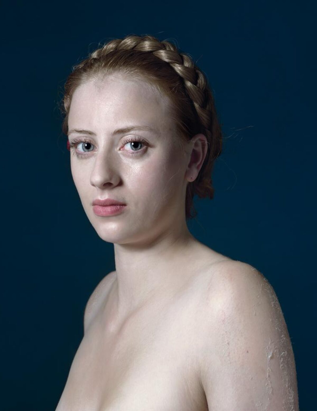 Portrait of a young woman with fair skin and braided hair styled close to her head, shown from the shoulders up against a deep teal background. She gazes toward the camera with a composed expression, her skin appearing wet or dewy, rendered in naturalistic detail with cool lighting that emphasizes the contrast between her pale complexion and the dark background.