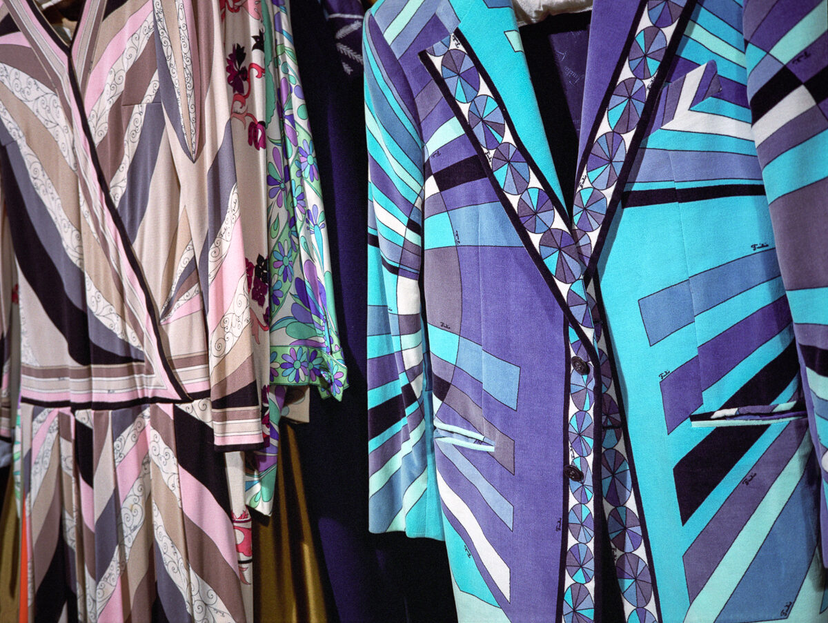 Colorful printed garments displayed on hangers, featuring bold geometric patterns and vibrant hues of turquoise, purple, pink, and cream. The designs incorporate repeating motifs including angular stripes, floral elements, and ornamental borders characteristic of mid-century fashion design.