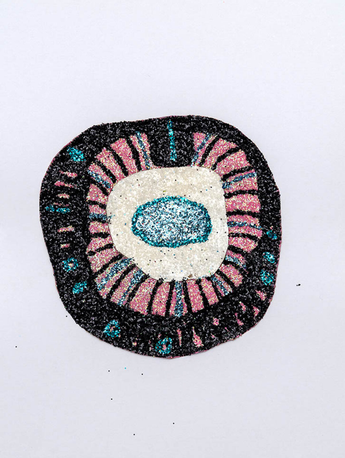 A concentric circular composition featuring a turquoise glitter eye at the center, surrounded by radiating pink and black glittered sections, with a cream-colored ring creating definition between layers. The iridescent glitter creates a shimmering, textured surface against a pale background.