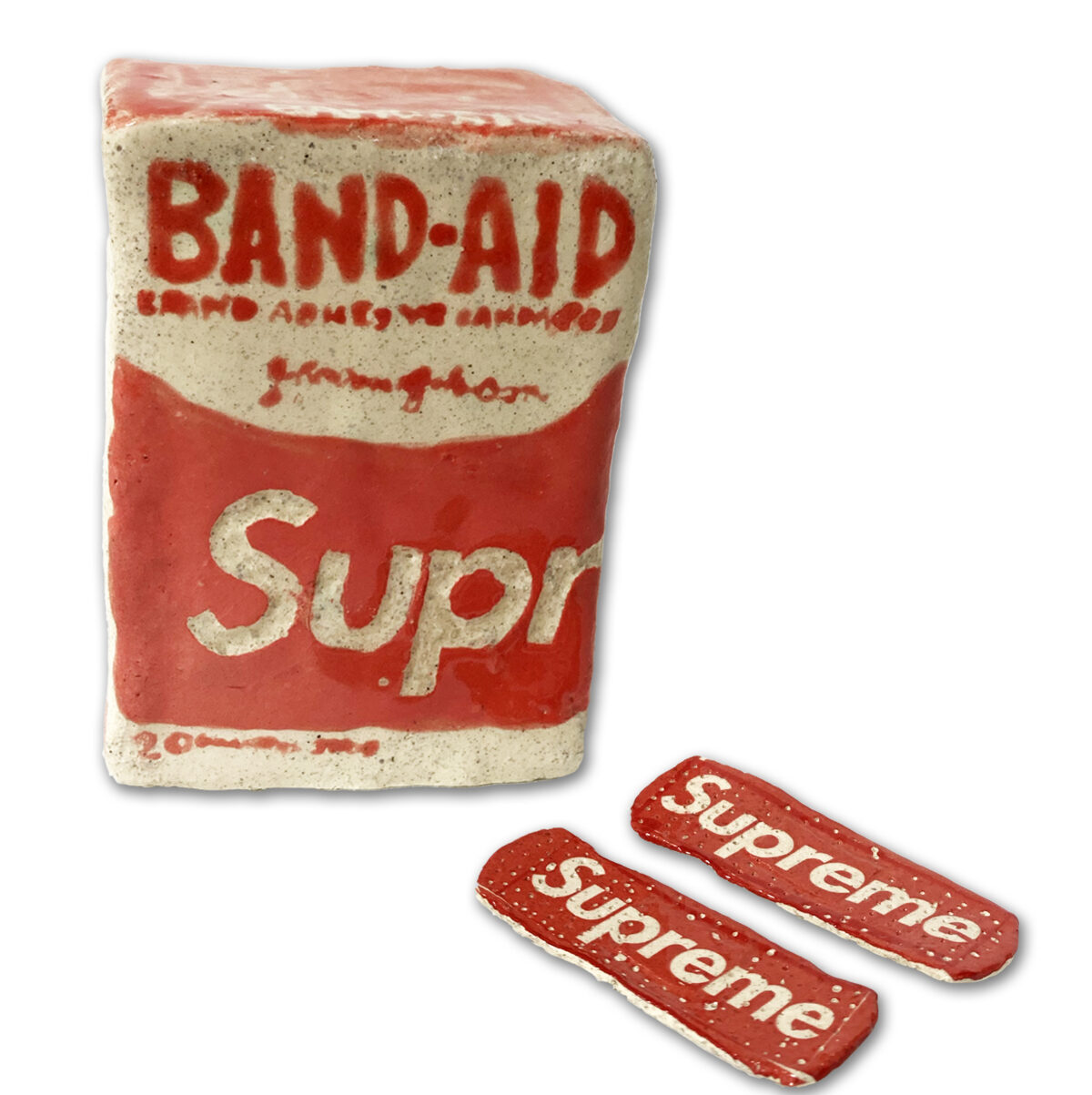 A ceramic sculpture of a vintage Band-Aid box rendered in cream and red glazed clay, accompanied by two small matching ceramic Band-Aid strips. The box features period typography and branding details in relief, with a weathered, aged appearance suggesting both the original product design and the materiality of handcrafted clay.