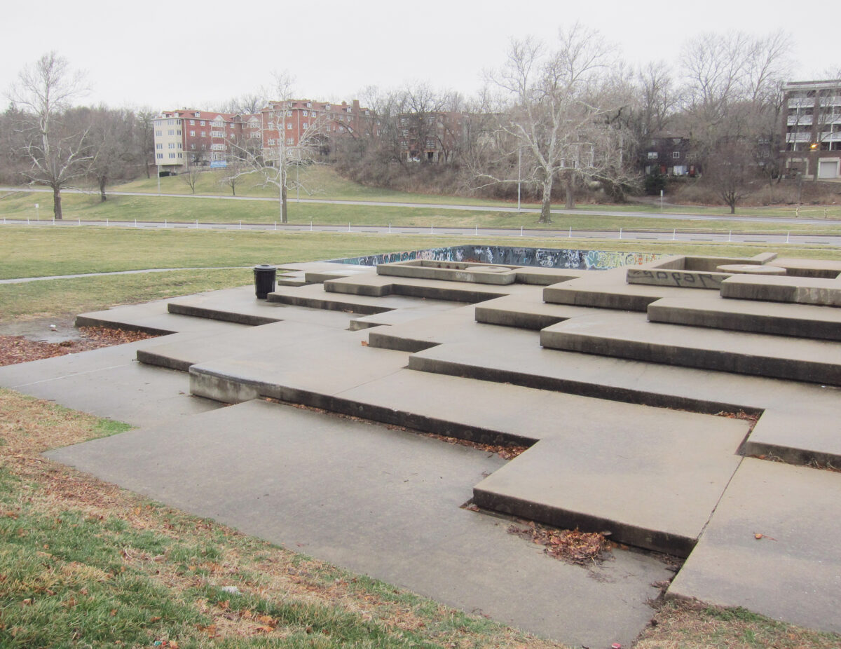 Concrete platforms and stepped forms of varying heights arranged across a paved plaza in an urban park, with bare trees and brick apartment buildings visible in the background under an overcast sky.