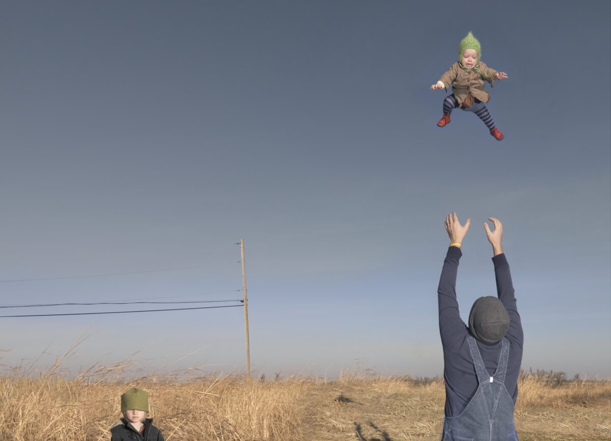 An adult wearing a dark jacket and green hat tosses a small child in a green knit hat high into a clear blue sky above a vast, dry grass field. Another child in a green hat stands in the foreground as the adult's arms reach upward to catch the airborne child, with a utility pole visible on the horizon.
