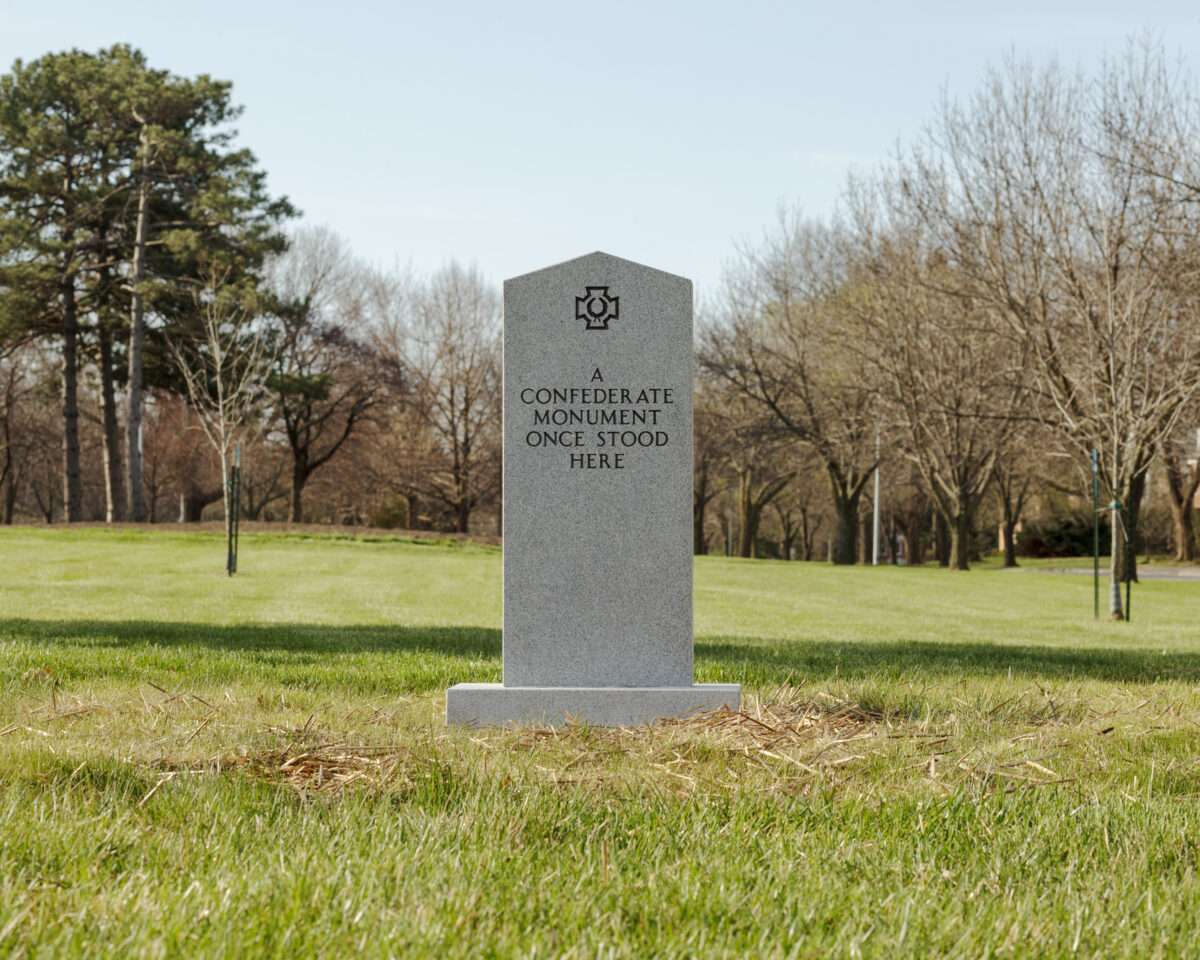 A gray stone monument stands alone in a grassy park surrounded by bare and evergreen trees. The obelisk-shaped marker bears an engraved symbol at its top and text reading "A CONFEDERATE MONUMENT ONCE STOOD HERE."