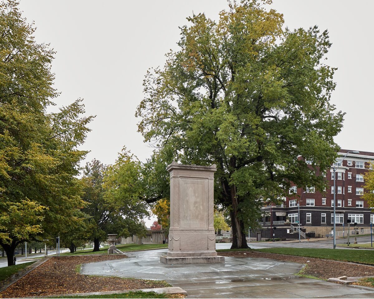 A wet stone monument pedestal stands centered in a public plaza, surrounded by mature trees in autumn foliage. Historic brick buildings frame the composition on the right, while tree-lined pathways extend into the distance on the left under an overcast sky.