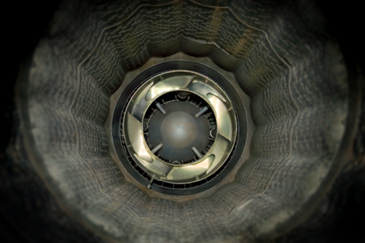 A symmetrical view down a cylindrical tunnel with concentric ribbed walls in gray and black tones, centered on a metallic circular aperture with reflective surfaces and a dark spherical core.