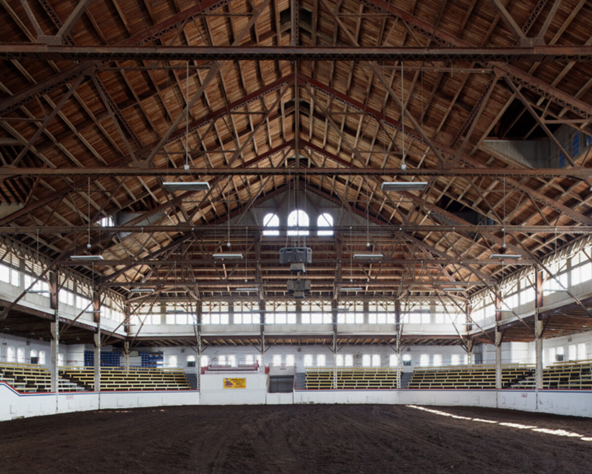 Interior view of a vast wooden-beamed arena with a dirt floor, yellow and white bleachers lining the sides, and natural light streaming through arched windows and skylights in the peaked roof structure.