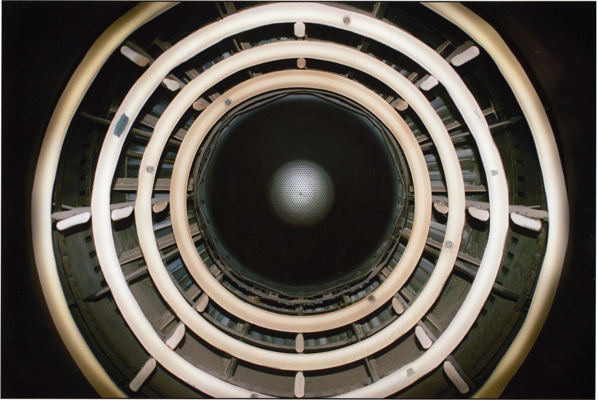 Concentric circles of cream and white rings with metal fixtures frame a dark central aperture, creating a geometric spiral effect. The composition suggests looking down into a cylindrical industrial or architectural structure, with alternating bands of light and shadow receding toward a barely visible illuminated center.