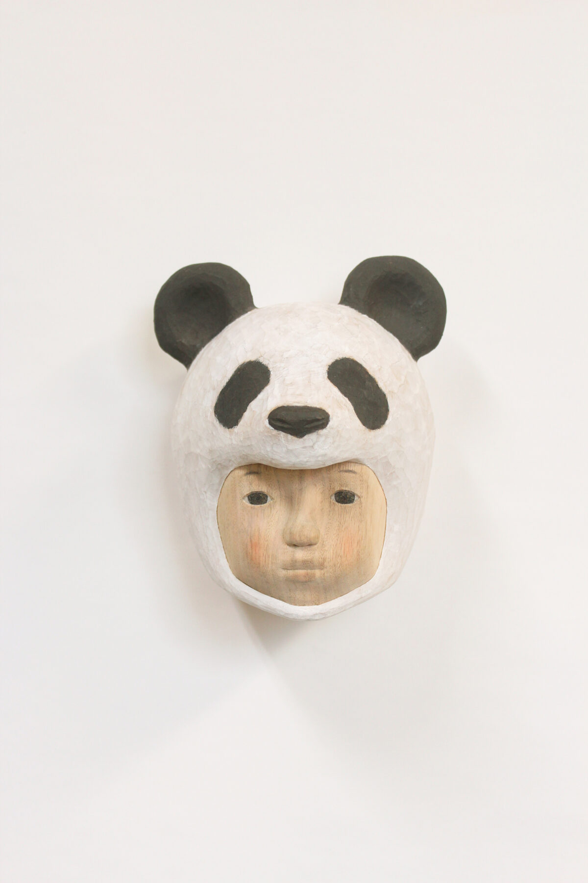 A carved wooden mask depicting a human face wearing a giant panda head covering, with black and white coloring and subtle flesh-toned facial features below. The sculpture combines naturalistic human portraiture with stylized animal characteristics, mounted on a white background.