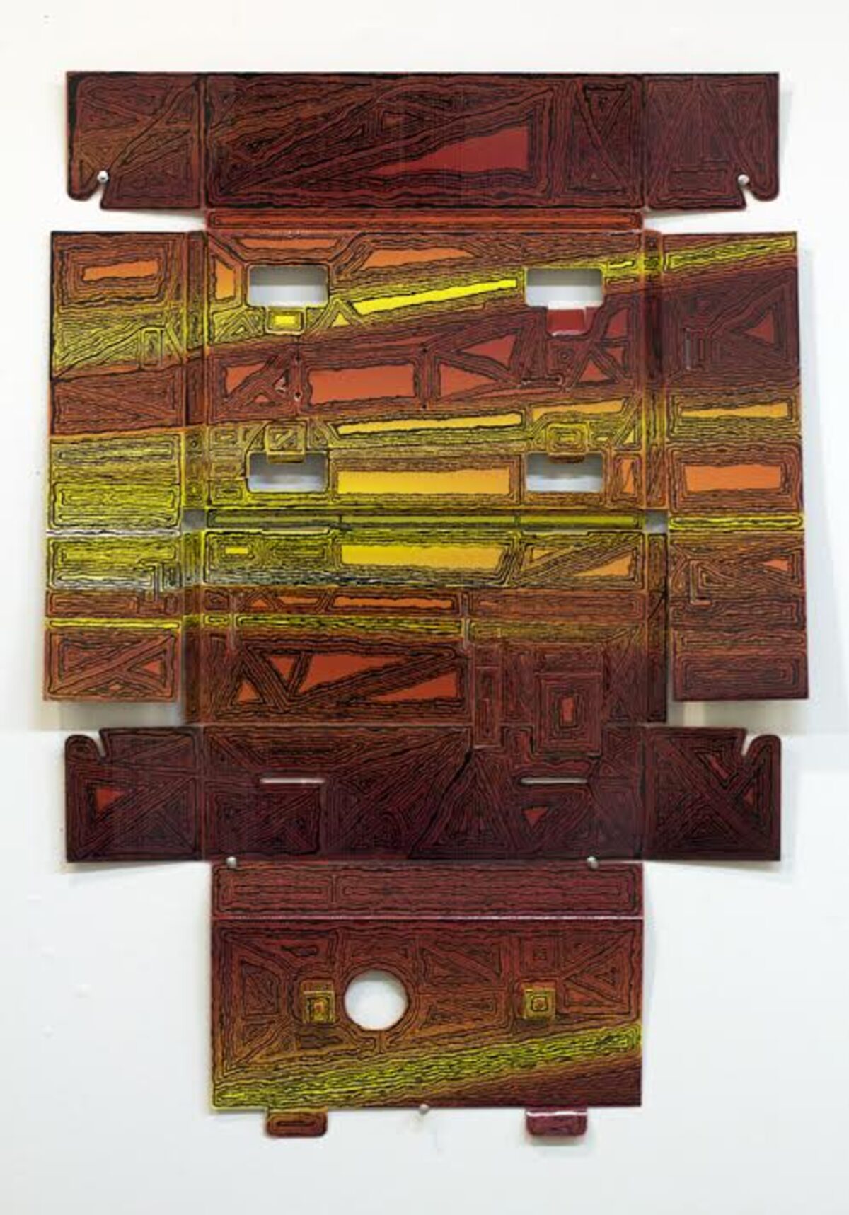 Abstract geometric composition made from layered cardboard pieces in warm earth tones—deep reds, browns, and yellows—with incised linear patterns creating a grid-like structure. White rectangular cutouts and openings punctuate the central composition, arranged in an asymmetrical, roughly symmetrical overall form against a white wall.