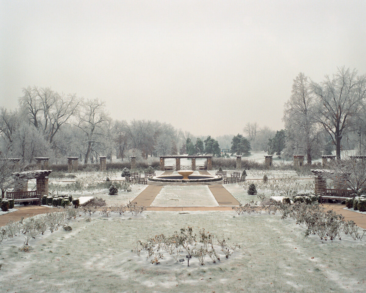 A frost-covered formal garden with a central circular fountain viewed down a symmetrical pathway. Bare trees frame the scene on both sides while brick structures and evergreens line the composition, all blanketed in ice and snow beneath an overcast sky.