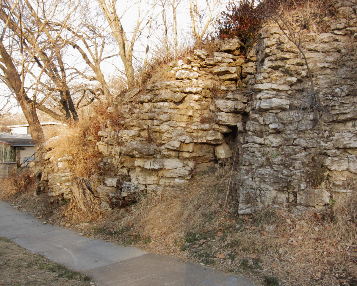 Stone ruins of stacked tan and grey limestone blocks form a tall wall alongside a concrete pathway, with bare winter trees and dried vegetation visible in the foreground and background.