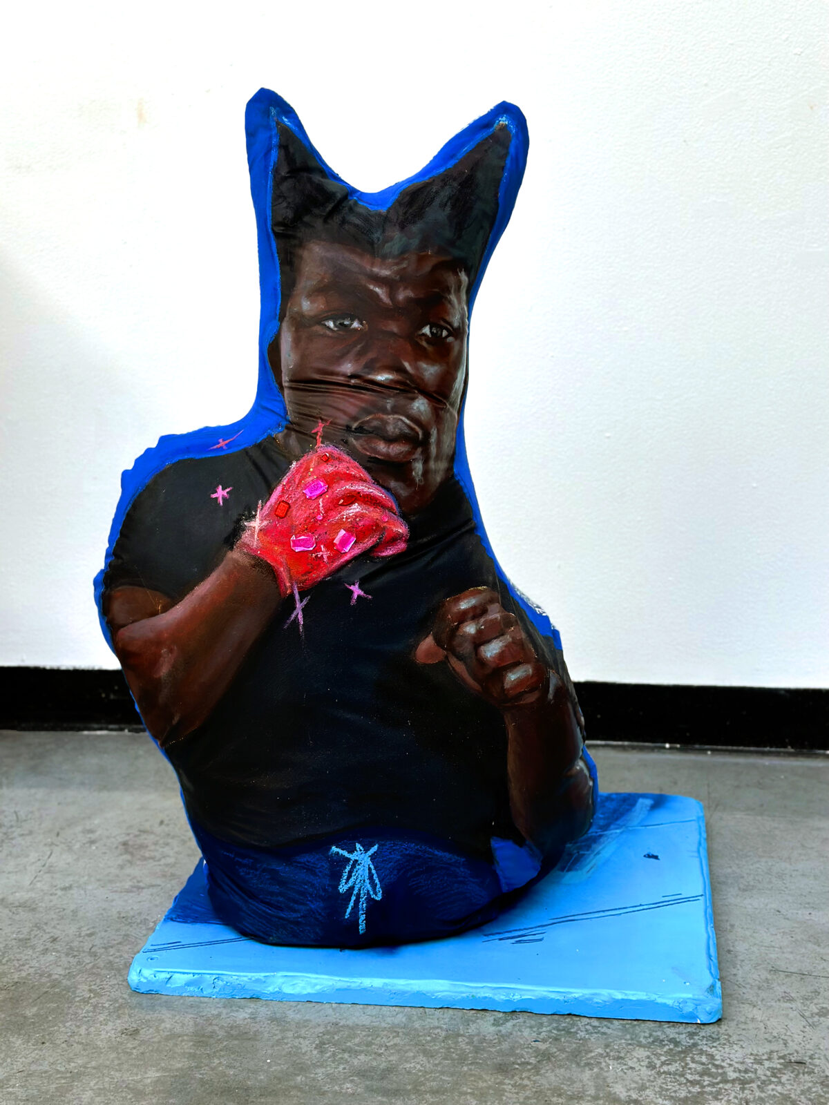 A sculptural figure with an exaggerated cat-like head in deep brown sits on a bright blue rectangular base. The figure wears a black shirt and blue pants, with one arm raised displaying a vivid red, textured glove adorned with pink embellishments. Small pink stars are scattered across the black garment, while a delicate blue figure sketch appears on the pants leg.