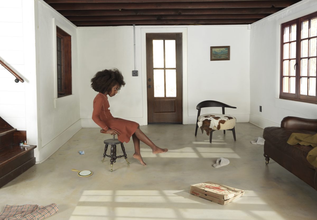 A woman in a rust-colored dress sits on a wooden stool in a sparsely furnished, sunlit room with concrete flooring, white walls, and dark wooden architectural elements. The minimalist interior contains scattered objects including chairs, a couch, pizza boxes, and dishes, creating an ambiguous domestic scene composed with careful attention to light and shadow.