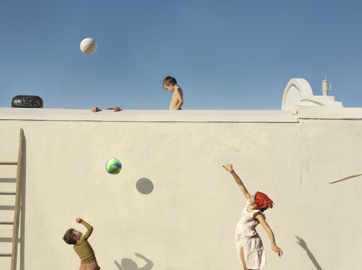 Children play with balls against a white concrete wall, with one child perched on top and others positioned below. The composition is divided between the bright blue sky above and the stark white architectural surface, creating a striking contrast that emphasizes the playful figures and their shadows cast on the wall.