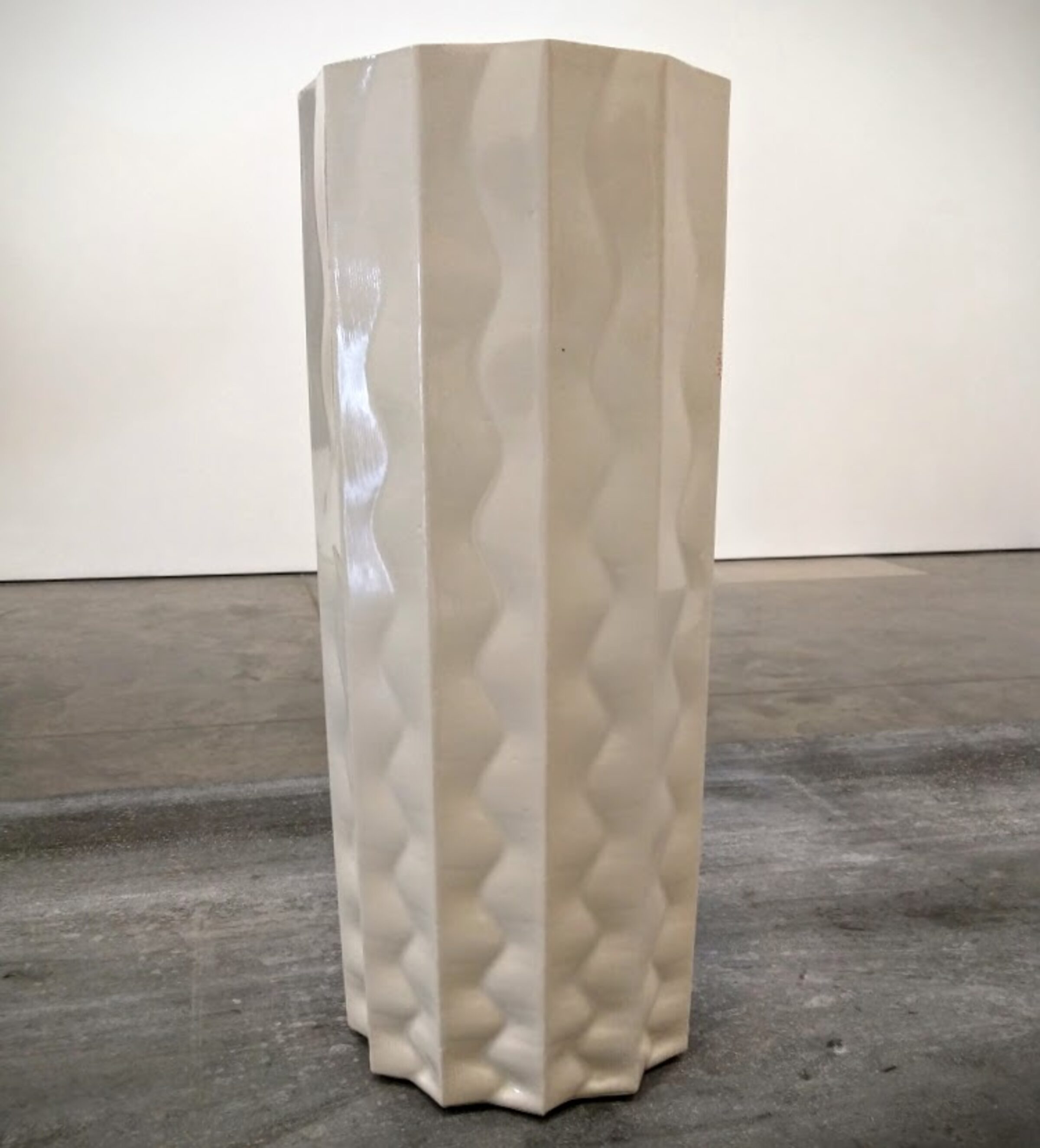 A tall, cylindrical white porcelain vase with a hexagonal cross-section stands on a concrete floor. The surface is decorated with repeating wavy vertical patterns in subtle relief, creating a quilted or undulating texture across all sides.