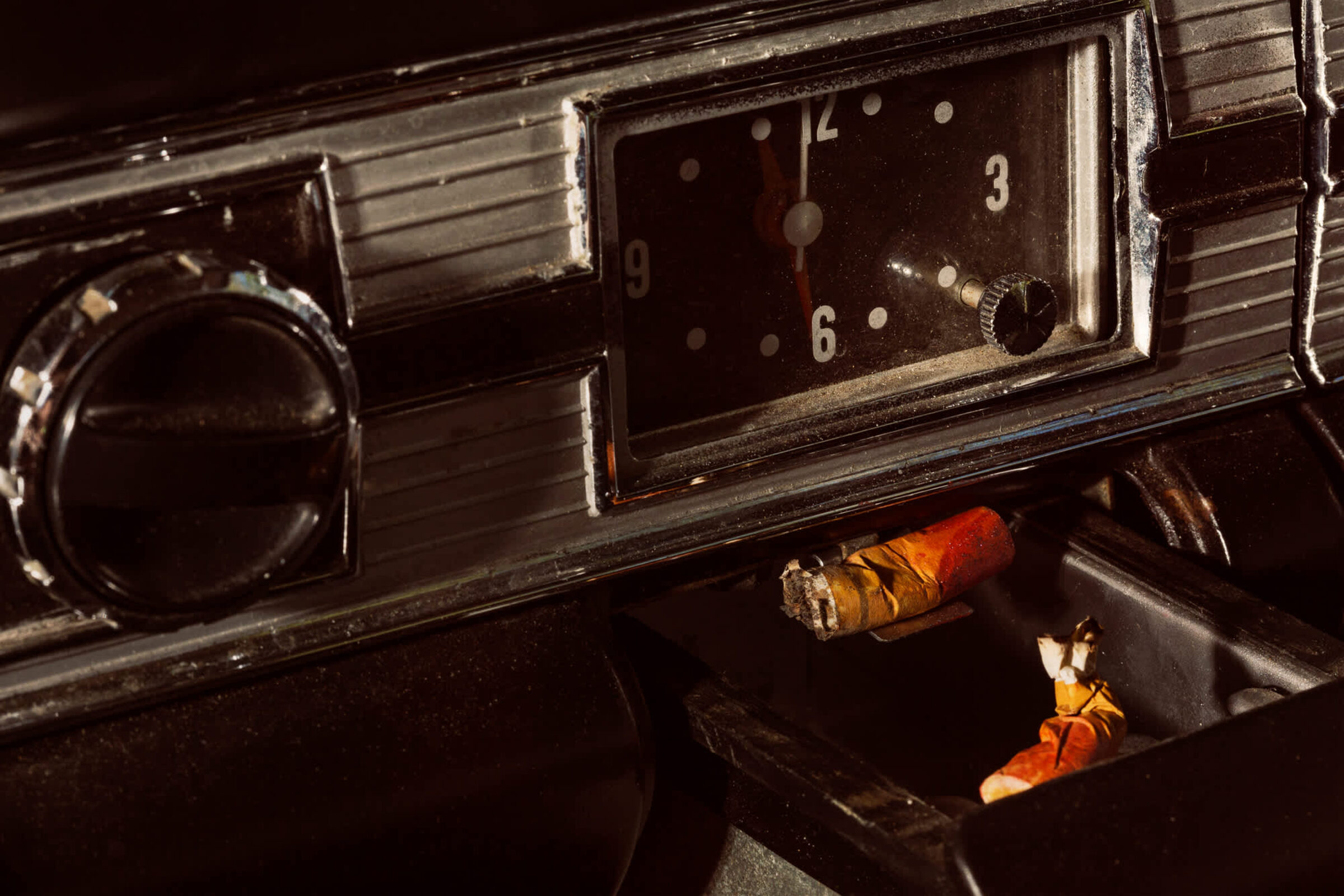 Vintage car dashboard shot from above in dramatic lighting, featuring a dark steering wheel on the left, an analog clock showing approximately 3:00, and a wooden cigar resting on the dashboard. Warm orange and amber tones of the cigar contrast sharply against the black interior and metallic dashboard trim.