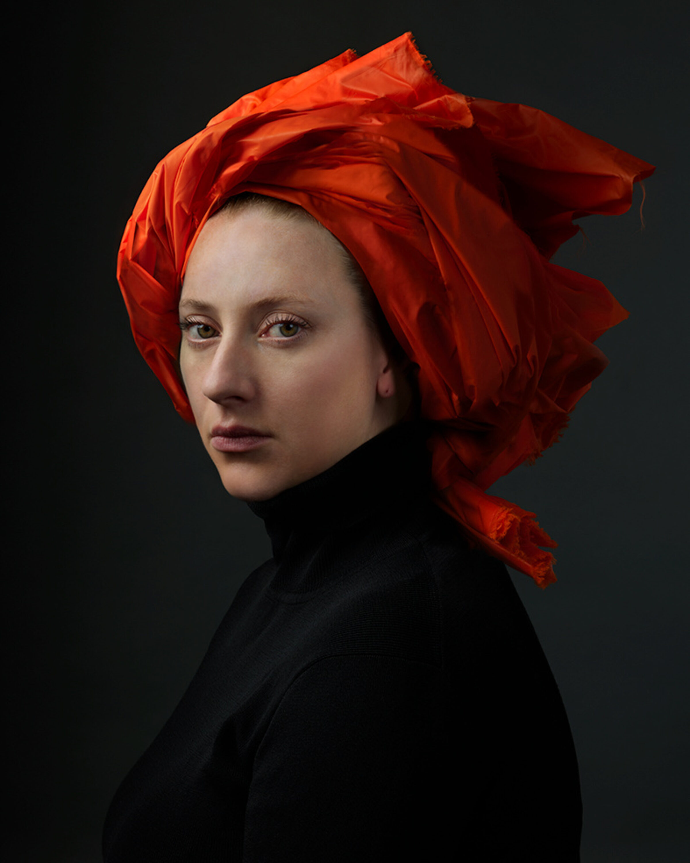 Portrait of a person with a pale complexion wearing a voluminous bright red-orange fabric wrapped around their head, set against a dark gray background. The figure is dressed in black and gazes toward the viewer in three-quarter profile, creating a striking contrast between the vivid headwrap and subdued tones of the clothing and background.
